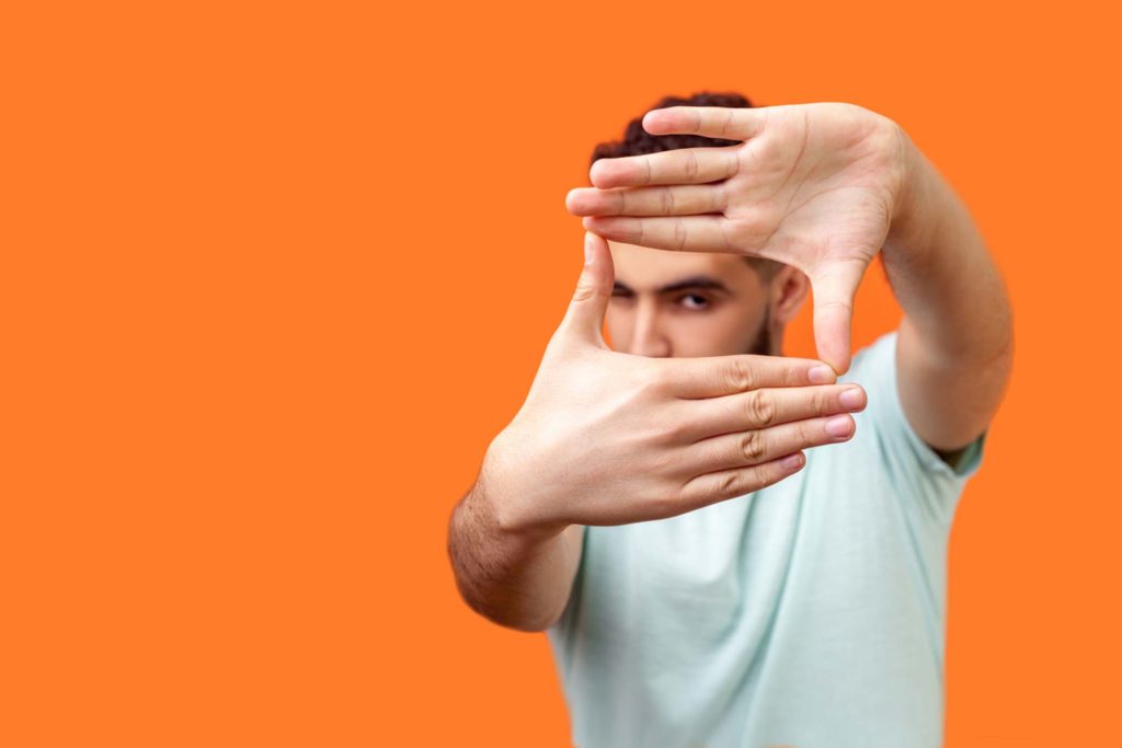 Portrait of attentive curious brunette man focusing through photo frame made of fingers. indoor studio shot isolated on orange background