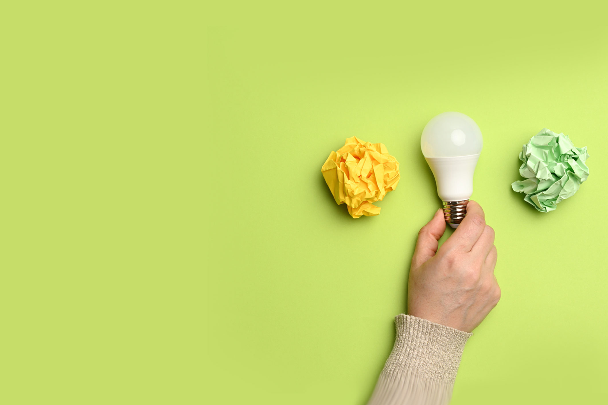 hand holding lightbulb between a crumbled piece of yellow paper and green paper - against lime green background