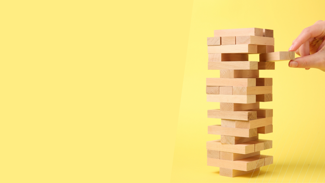 Hand carefully removing a wooden block from a stacked tower, representing the strategic planning, risk management, market validation, and cross-functional coordination required for a successful new product introduction in B2B technology markets.